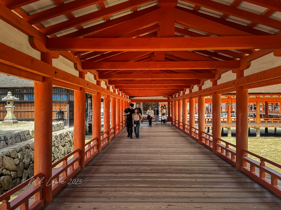 A wooden walkway with red beams and support columns, leading into a traditional Japanese structure, with people walking along it.