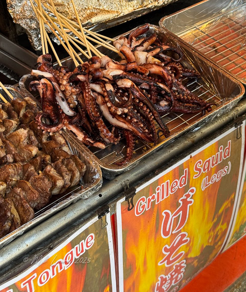 A close-up view of skewered grilled octopus legs being sold at a street food stall, with a sign reading 'Grilled Squid (Legs)' in Japanese.