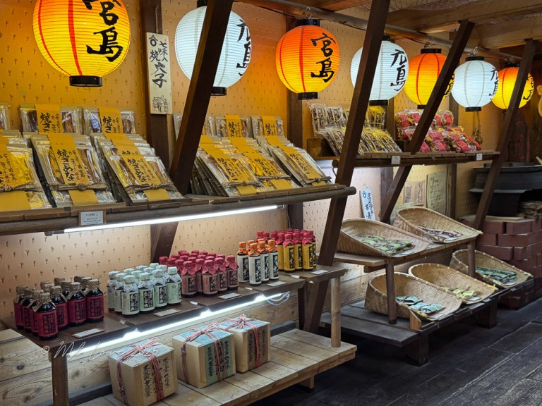 Interior view of a shop in Miyajima Island featuring local products, including colorful bottles and packaged snacks, with traditional lanterns hanging above.