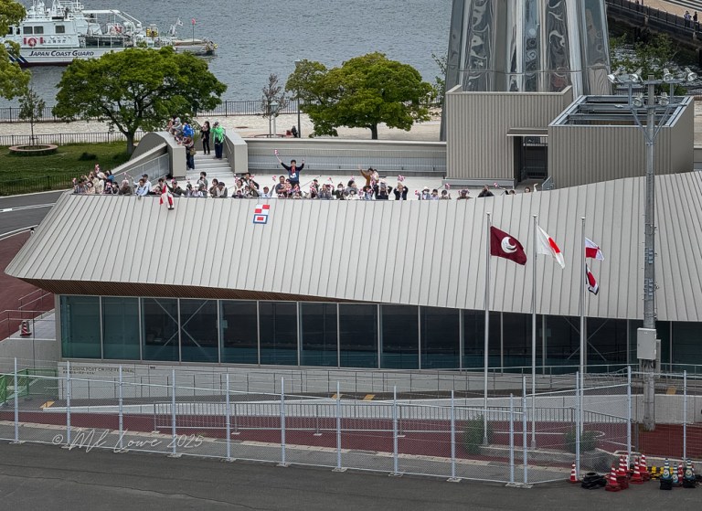 A crowd of people waving from a modern building in Hiroshima, surrounded by greenery and water, with various flags displayed.