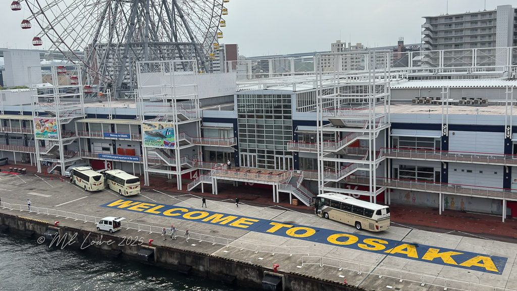 A view of the Osaka port area featuring large buses parked beside a building, with a prominent 'WELCOME TO OSAKA' painted on the ground and a ferris wheel in the background.