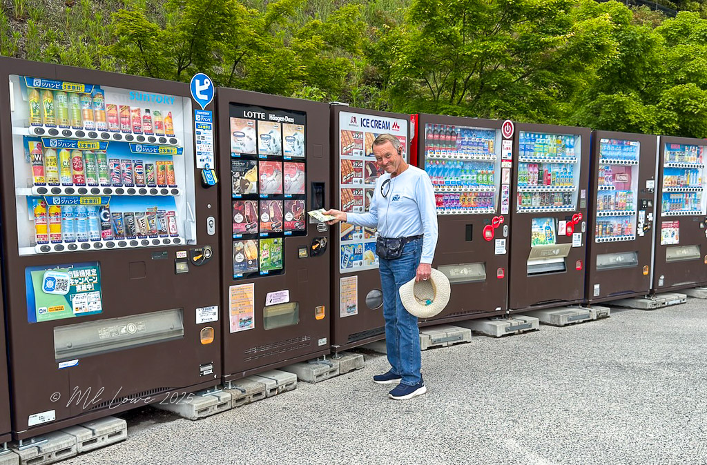 A man stands in front of multiple vending machines in Japan, selecting a beverage. The machines feature various drinks, snacks, and ice cream options.