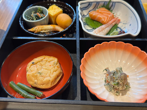 A traditional Japanese-style bento box featuring assorted dishes including salmon sashimi, tempura, a sweet egg omelette, green beans, and other side items.