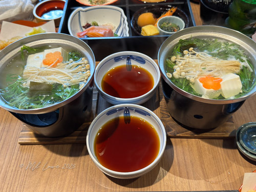 A Japanese hot pot dish with two bowls of clear broth containing vegetables, tofu, and mushrooms, accompanied by three smaller bowls of dipping sauce, set on a wooden table.