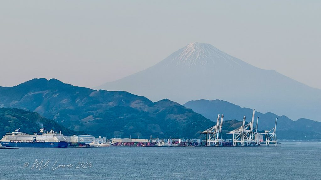 A scenic view of Shimizu, Japan, featuring the coastline with ships and cranes in the foreground, while the iconic Mt. Fuji is visible in the background, partially shrouded in mist.