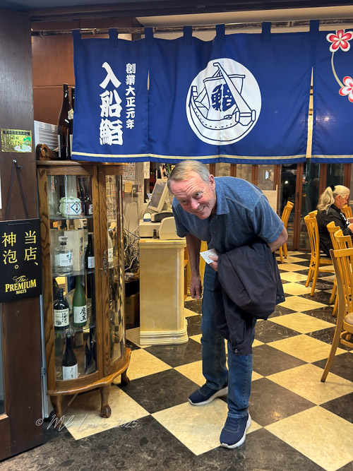 A man standing at the entrance of a restaurant with a blue fabric sign in Japanese above him, showcasing various bottles of sake and wooden chairs visible in the background.