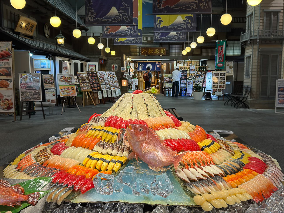 A vibrant display of sushi and sashimi in a market setting, featuring colorful sushi pieces arranged artistically on a large platter, with menus and signs from nearby restaurants in the background.