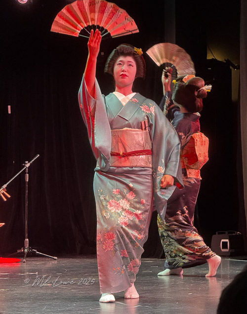 A geisha performing on stage, dressed in a traditional kimono, holding a fan, with another performer in the background.