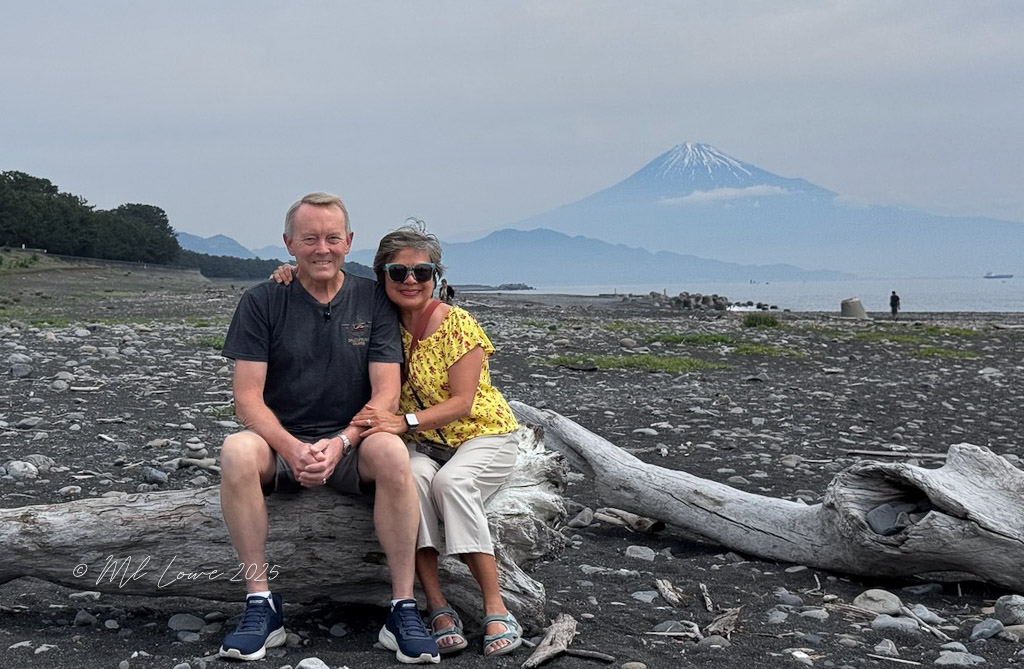A couple sitting on a log at a beach with a view of Mount Fuji in the background, wearing casual clothing and sunglasses, with a rocky shoreline and scattered vegetation.