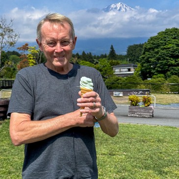 A person holding an ice cream cone with Mount Fuji in the background, surrounded by greenery and a cloudy sky.