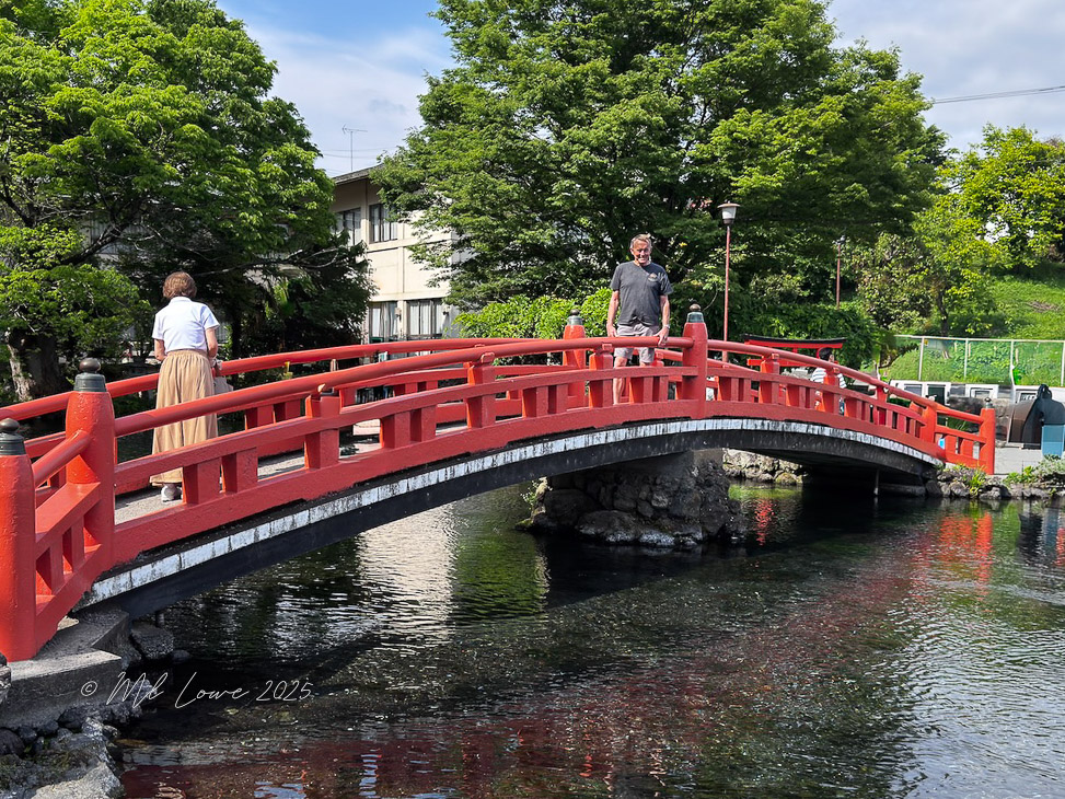 Two people standing on a red bridge over clear water, surrounded by greenery.