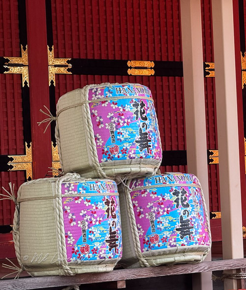 Three traditional sake barrels with colorful labels, stacked in front of a decorative wooden wall.