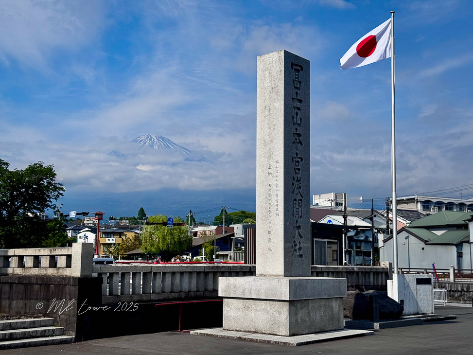 A stone monument with inscriptions stands in the foreground, while a Japanese flag flutters nearby. In the background, Mount Fuji is partially obscured by clouds and a blue sky.