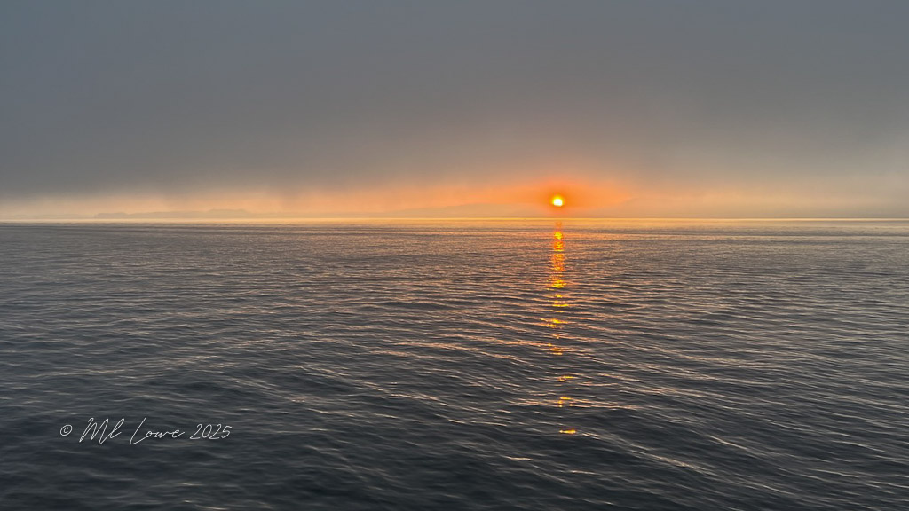 A serene sunset over a calm sea, with the sun's reflection glistening on the water's surface under a misty sky.