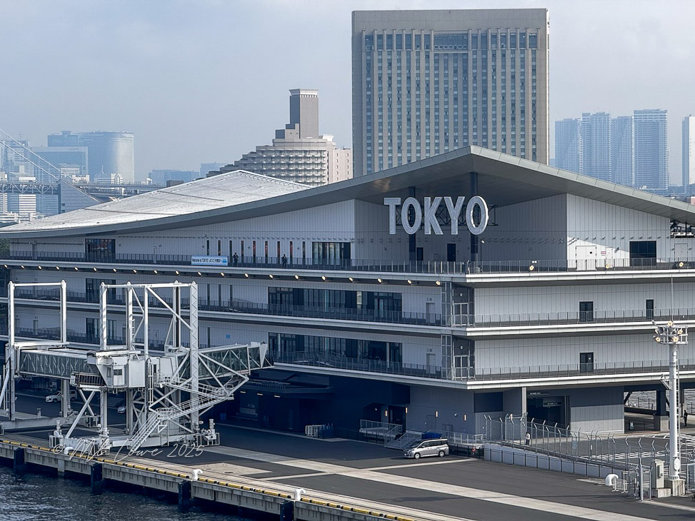 The exterior of a modern building with a large 'TOKYO' sign on the roof, featuring multiple stories and a sleek design, situated along a waterfront in an urban setting.