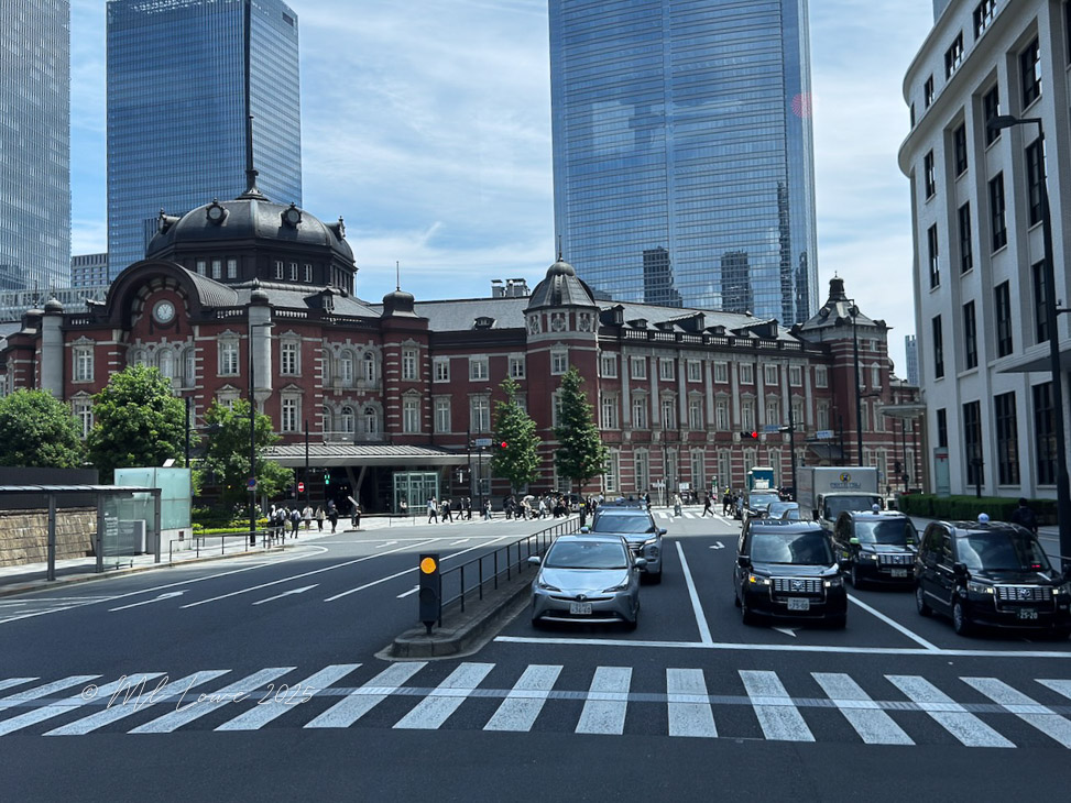 View of Tokyo Station, a historical red-brick building, surrounded by modern skyscrapers. The scene includes a busy street with cars and pedestrians crossing at a crosswalk.