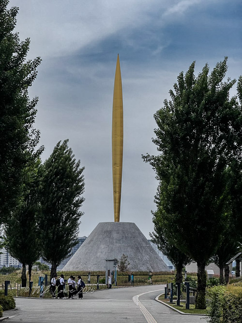 A golden needle-like monument atop a gray pyramid structure, surrounded by lush green trees, with a group of people gathered nearby.