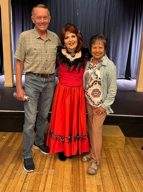 Three people posing together on stage after a performance, with one wearing a vibrant red dress and the others in casual attire.