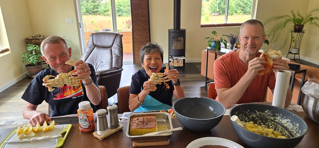 Three people sitting at a dining table, each holding a Dungeness crab, with food items and condiments in view, and a cozy interior background.