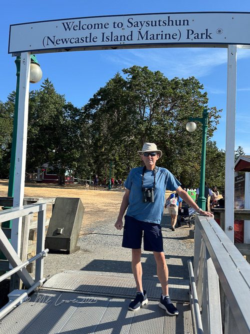 A person standing on a dock at Newcastle Island Marine Park, wearing a hat and binoculars, with a sign welcoming visitors in the background.