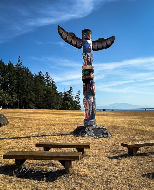 A vibrant totem pole with outstretched wings stands prominently in a field, surrounded by trees and overlooking a body of water.
