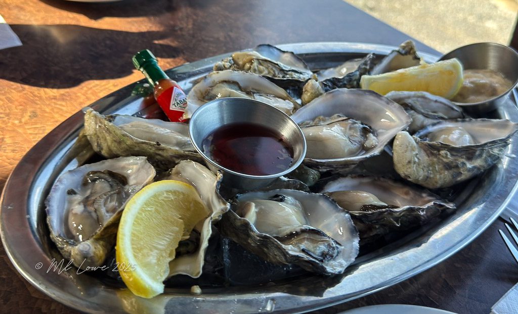 A metal platter filled with freshly shucked oysters, accompanied by lemon wedges, cocktail sauce, and a bottle of hot sauce.