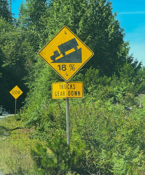 Road sign indicating an 18% grade with a warning for trucks to gear down, surrounded by trees and greenery.