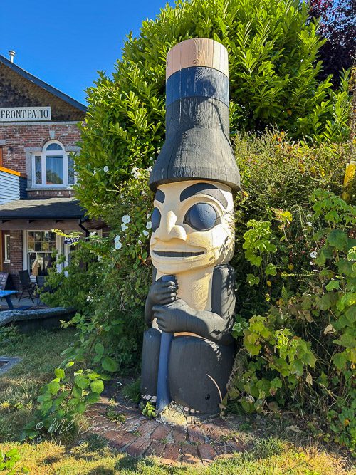 A carved wooden totem pole figure wearing a top hat and seated amidst greenery near a building.