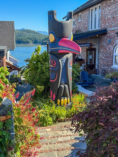 A colorful totem pole standing in a garden with lush greenery, stone steps leading up, and a view of the water and mountains in the background.