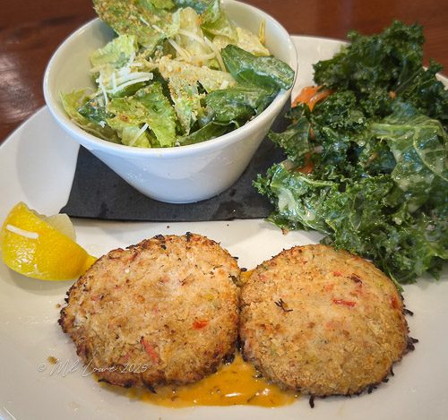 A plate featuring two crab cakes served with a side of Caesar salad and a fresh green salad, garnished with a lemon wedge.