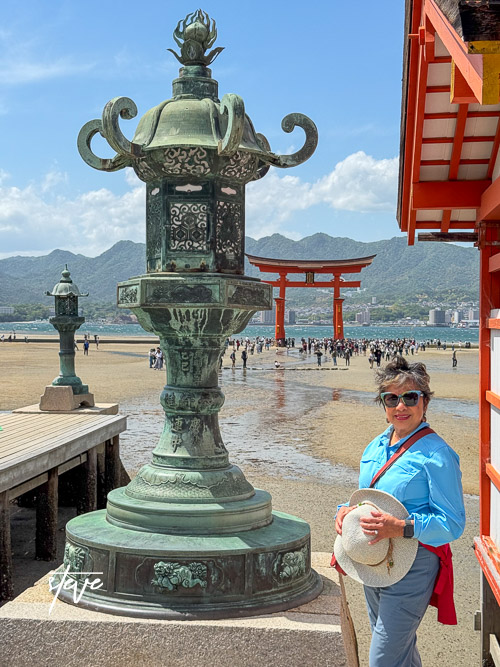 A person stands next to a decorative lantern near the Itsukushima Shrine's Torii Gate on Miyajima Island, with tourists and mountains in the background.