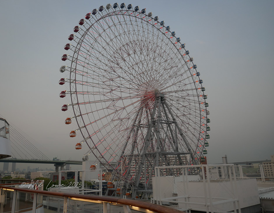 A large Ferris wheel with colorful cabins is prominently displayed against a soft evening sky in Osaka, Japan.