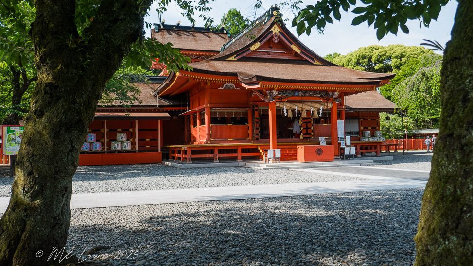 A traditional Japanese shrine building with vibrant red paint, set among trees and a gravel path, showcasing the architectural style characteristic of Shinto shrines.