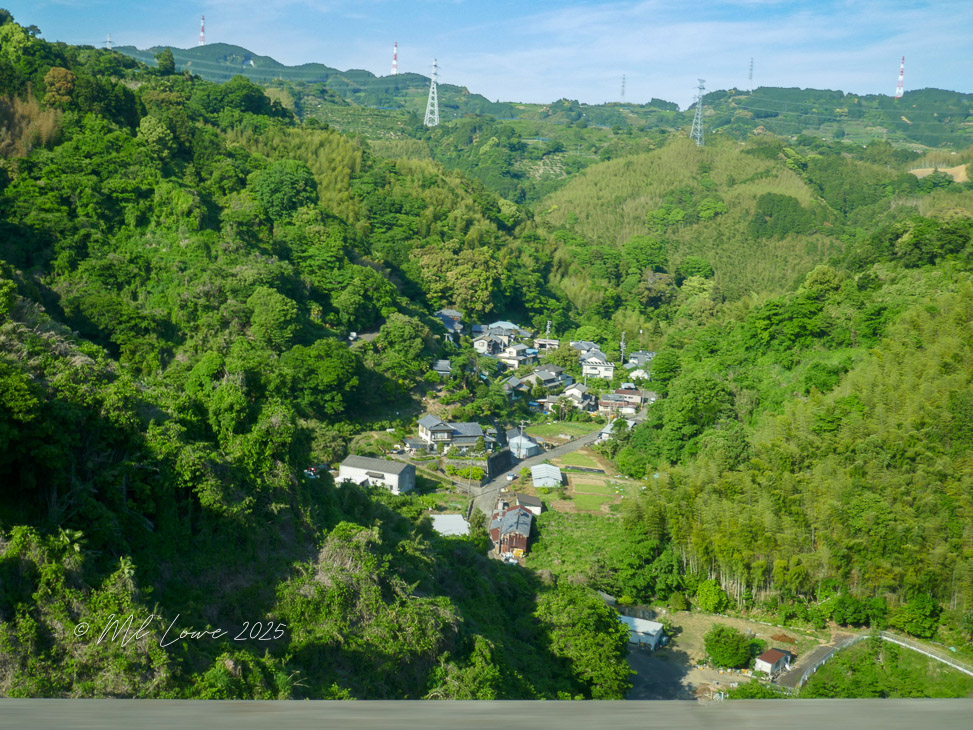 A scenic view of a rural Japanese village surrounded by lush greenery and hills, showcasing traditional homes amidst a mountainous landscape.