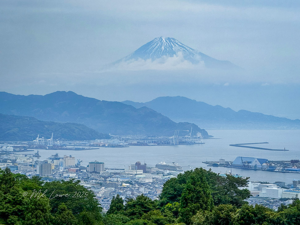 A hazy view of Mount Fuji rising above Shimizu City and Suruga Bay, showcasing a mix of urban and natural landscapes.