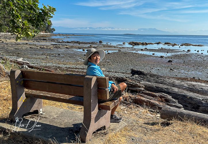 A person sitting on a wooden bench by the shore, smiling while facing the ocean with rocky land and distant mountains in the background.