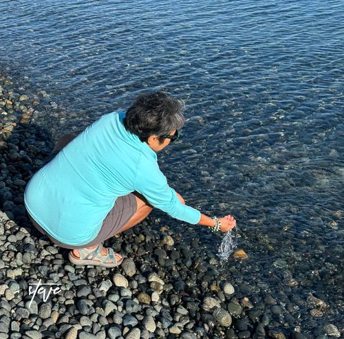 A person in a blue shirt kneeling by the water's edge, gently reaching to touch the clear water while surrounded by smooth pebbles.