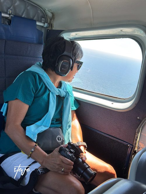 Person wearing headphones and holding a camera, looking out the window of a small aircraft.