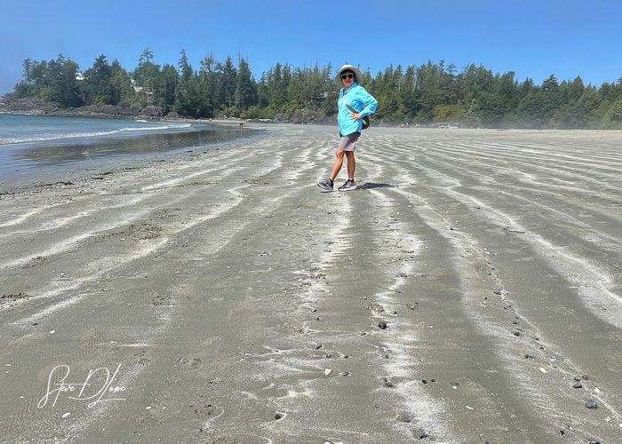 Person standing on a sandy beach with patterned sand and trees in the background on a sunny day.