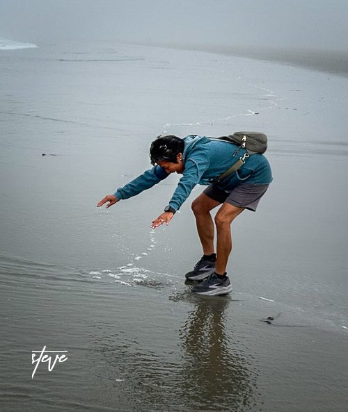 Person bending down to touch the water on a foggy beach.