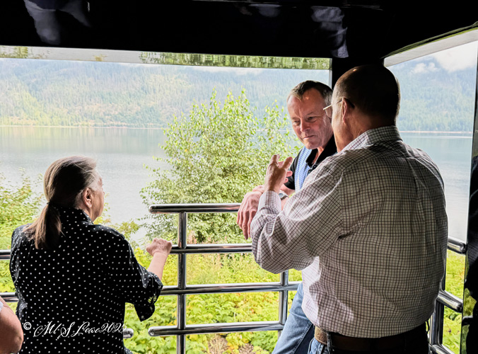 Three people interacting and talking in a scenic train cabin, with greenery and water visible through the windows.