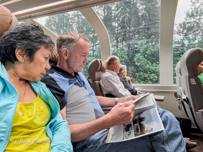 Two passengers inside the Rocky Mountaineer train, one reading a brochure and the other looking out the window, with fellow travelers visible in the background.