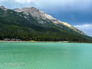 A serene view of a turquoise lake surrounded by vibrant green forests and mountains under a partly cloudy sky.