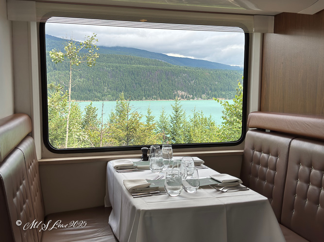 Interior view of a dining area on the Rocky Mountaineer train, featuring a table set with glassware and silverware, overlooking lush greenery and a turquoise lake through a large window.