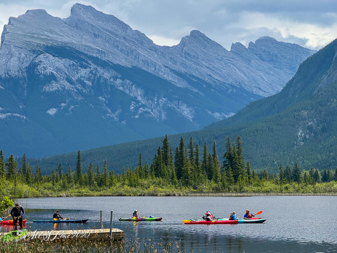 A group of kayakers paddling on a tranquil lake surrounded by lush forests and majestic mountains in the background.