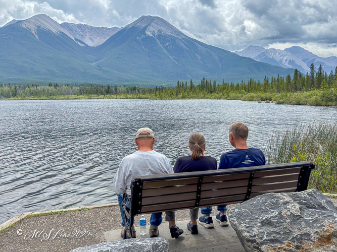 Three people sitting on a bench overlooking a calm lake surrounded by mountains and forest, with a cloudy sky above.