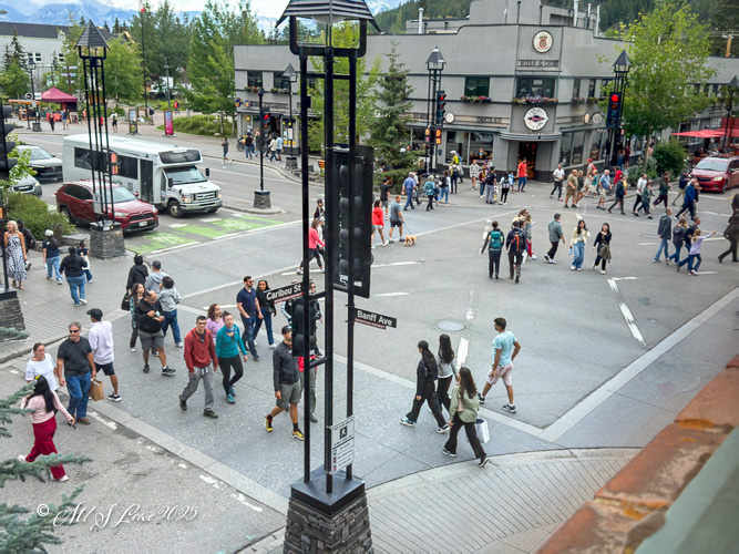 A busy intersection in the town of Banff, Alberta, featuring pedestrians crossing the streets and vehicles waiting at traffic signals, with local shops and trees in the background.