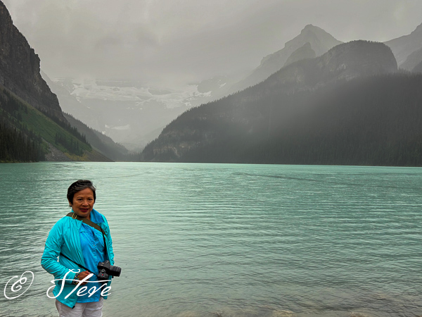 A person standing by a turquoise lake surrounded by mountains under overcast skies.
