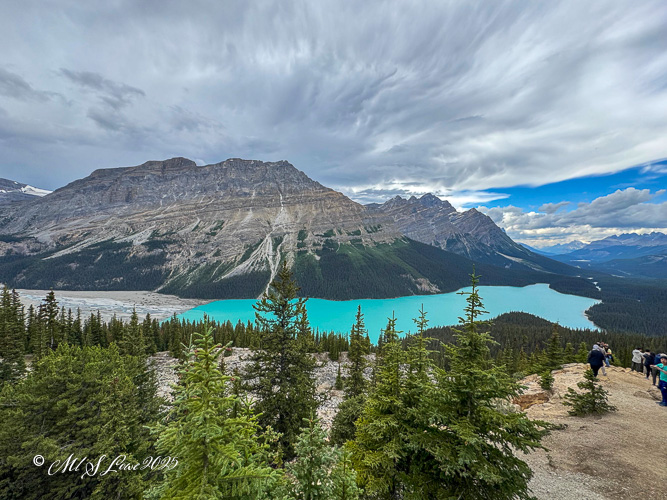 A breathtaking view of a turquoise lake surrounded by towering mountains and lush green trees under a dramatic cloudy sky, with visitors admiring the scenery.