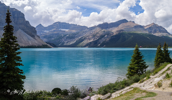 A panoramic view of a turquoise lake surrounded by majestic mountains and lush greenery, under a partly cloudy sky.